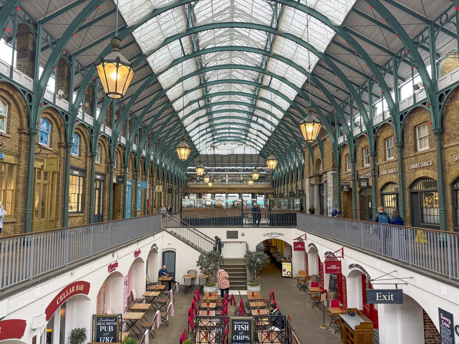 A vaulted indoor marketplace features arched walkways, shopfronts, and outdoor dining areas with people scattered throughout.